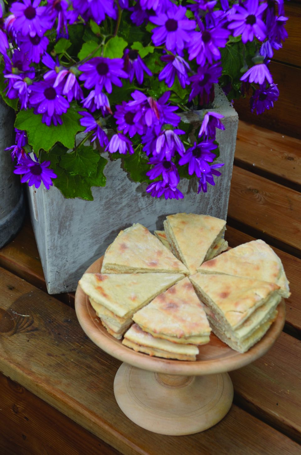 Lefse skåret i trekanter på et kakefat av tre, foran en krukke med lilla blomster